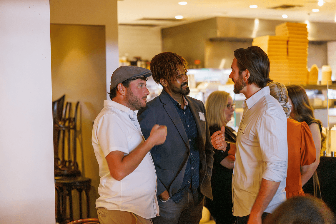 Three men, two wearing name tags, are engaged in conversation at an indoor event, with other attendees and a restaurant kitchen visible in the background.