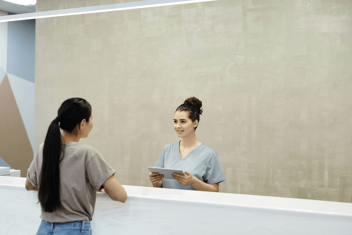 A smiling medical professional in scrubs holds a tablet, interacting with a patient at a modern reception desk.