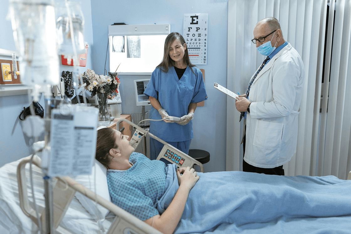 A nurse in blue scrubs and a doctor in a lab coat and mask consult with a smiling female patient lying in a hospital bed.