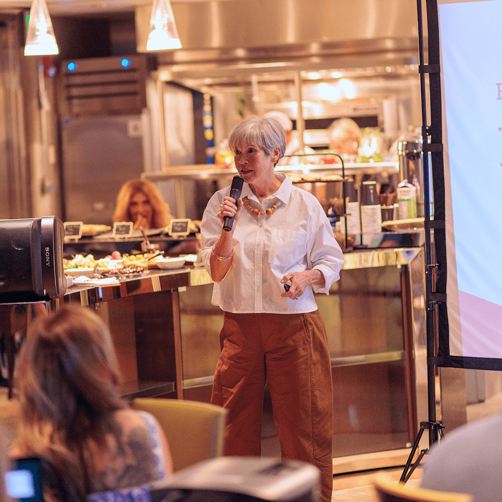 Older woman with grey hair speaking into a microphone at a presentation, with a food buffet and screen in the background.