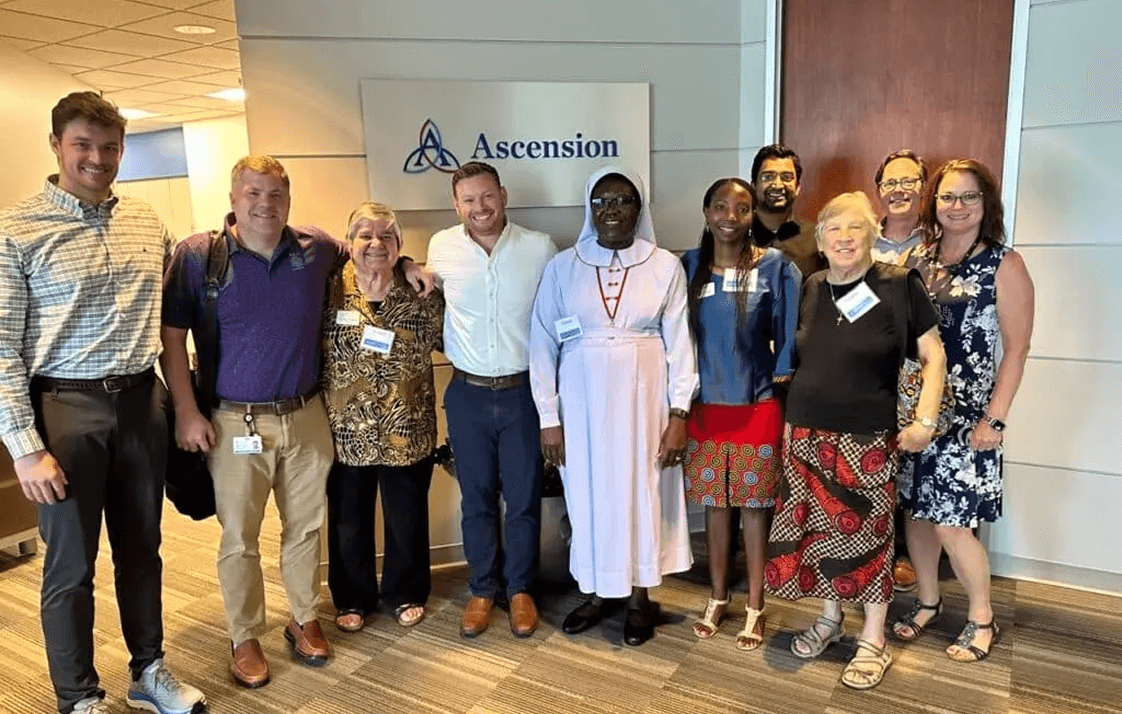 A diverse group of eleven smiling individuals, including a nun, stands in an office in front of an Ascension logo.
