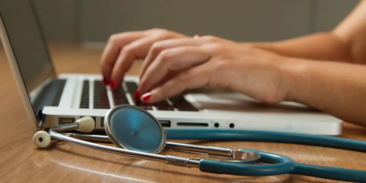 A medical professional types on a laptop, with a blue stethoscope on a wooden desk, symbolizing digital healthcare or medical administration.