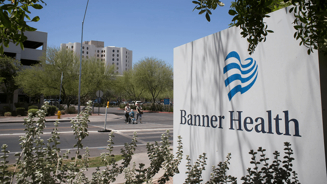 A prominent white Banner Health sign with its blue heart logo, situated at the entrance to a hospital campus on a sunny day.