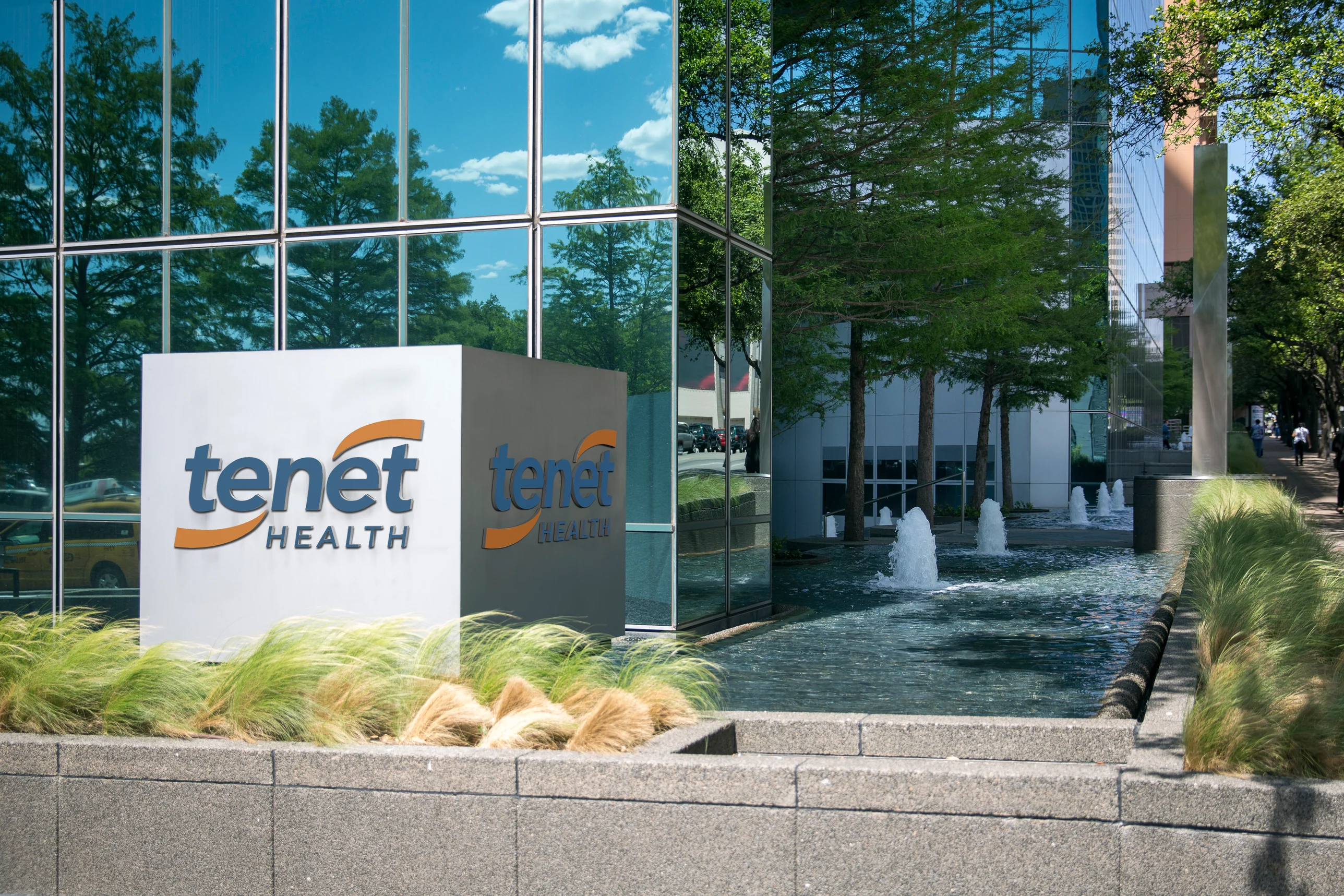 Tenet Health sign in front of a modern glass building reflecting trees and sky, alongside water fountains and lush grass.