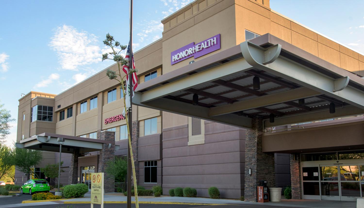 Modern HonorHealth hospital building with beige and brown facade, featuring a prominent purple "HONORHEALTH" sign, a red "EMERGENCY" entrance, and large covered walkways.