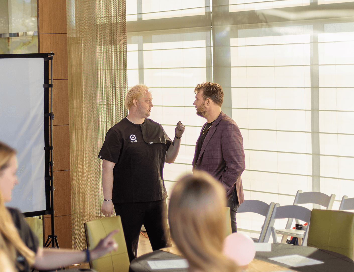 Two men, one in black scrubs and another in a blazer, converse at an event, with a projection screen and blurred attendees in the foreground.