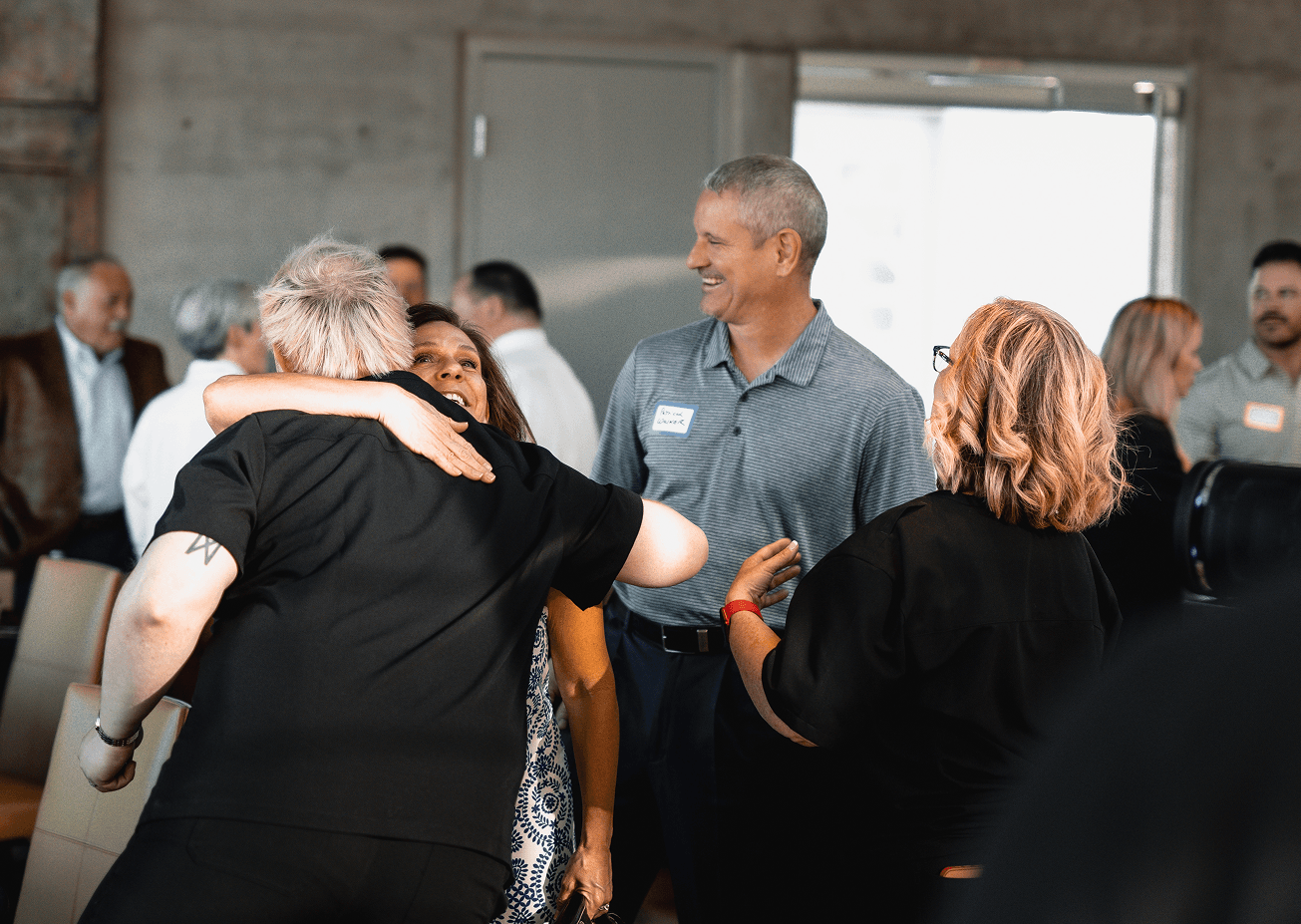 Two people hug warmly at a social event while a man with a nametag smiles nearby and another woman watches.