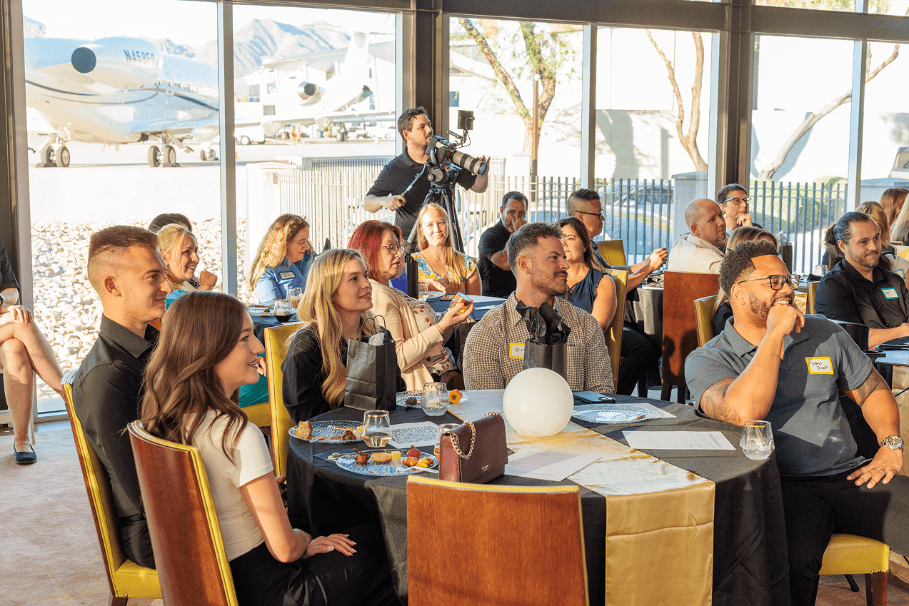 People socialize and listen at an indoor event, seated at tables with refreshments. A cameraman records, and private jets are visible outside through large windows.