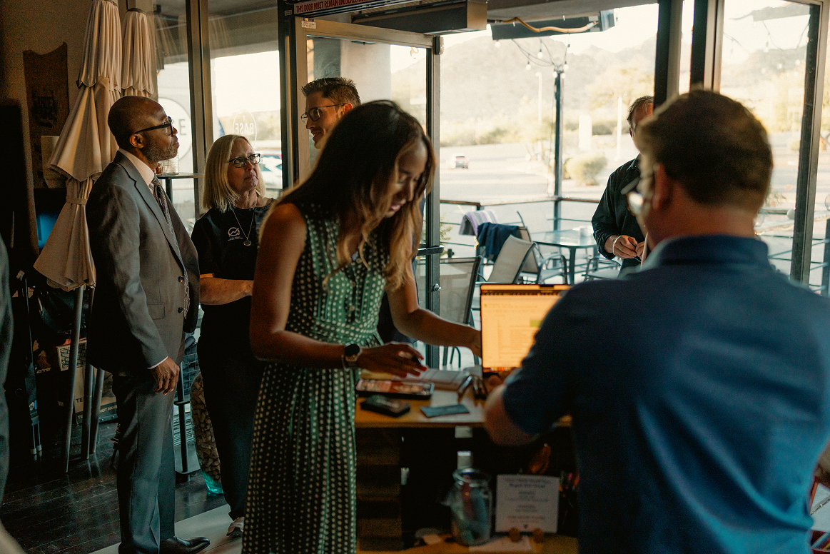 People gather inside a modern cafe or business, with a woman in a green polka dot dress interacting at a counter with a laptop as others observe.