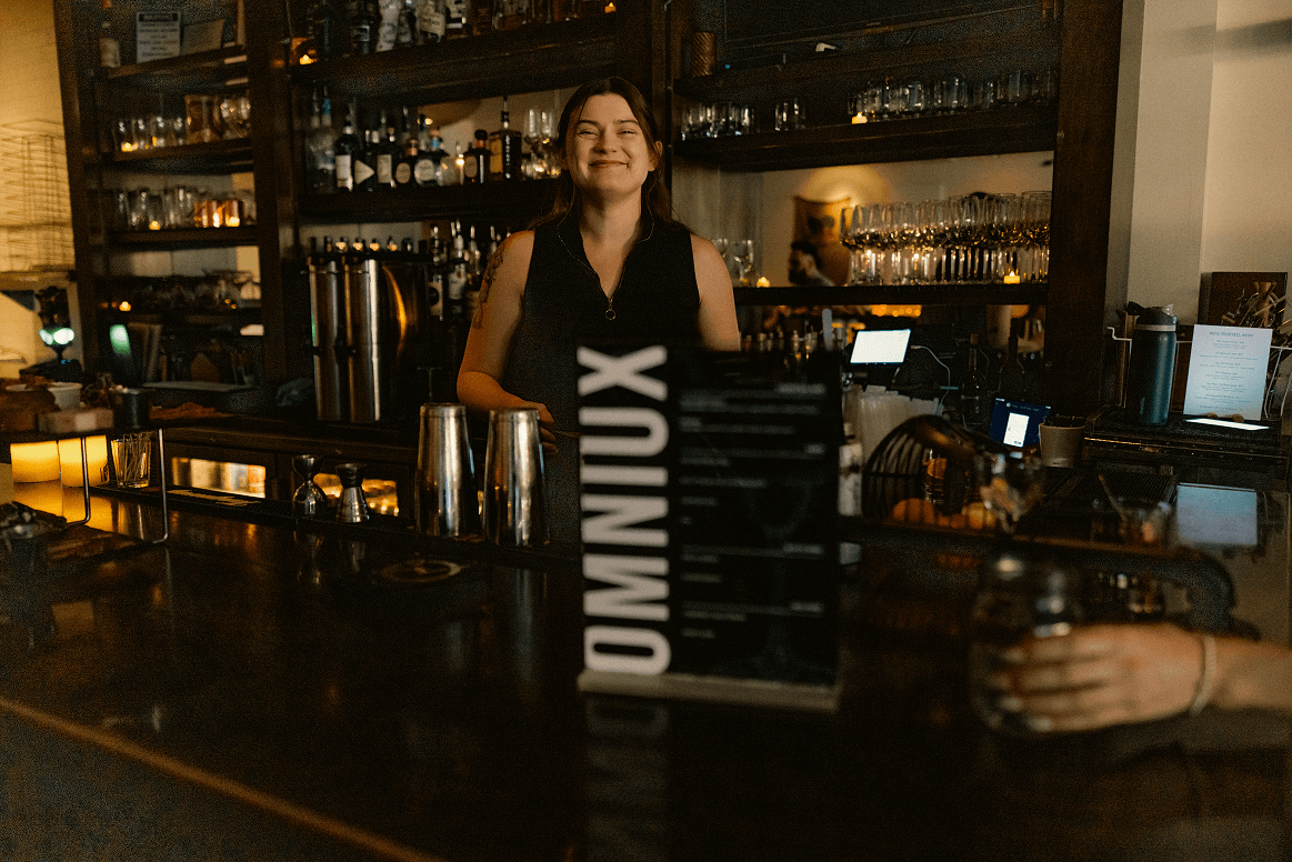 A smiling bartender stands behind a dark wooden bar, with shelves of bottles and glasses in the background. An "OMNIUX" sign is on the bar, and a customer's hand holds a drink in the foreground.