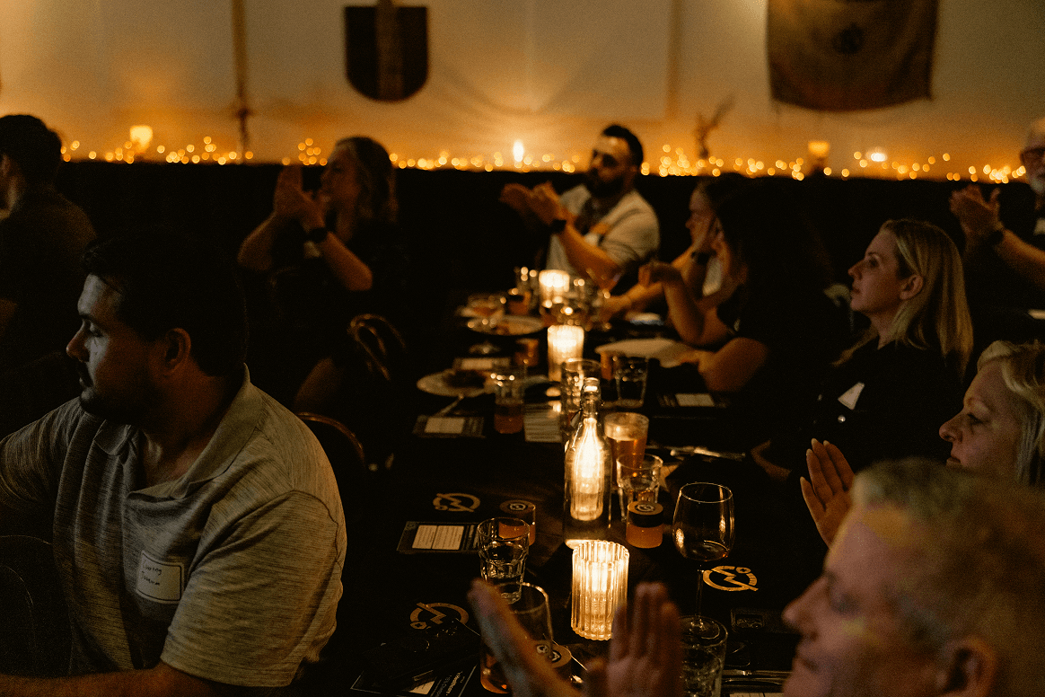 Attendees at a dimly lit indoor evening event, illuminated by candles and string lights, with several people clapping.