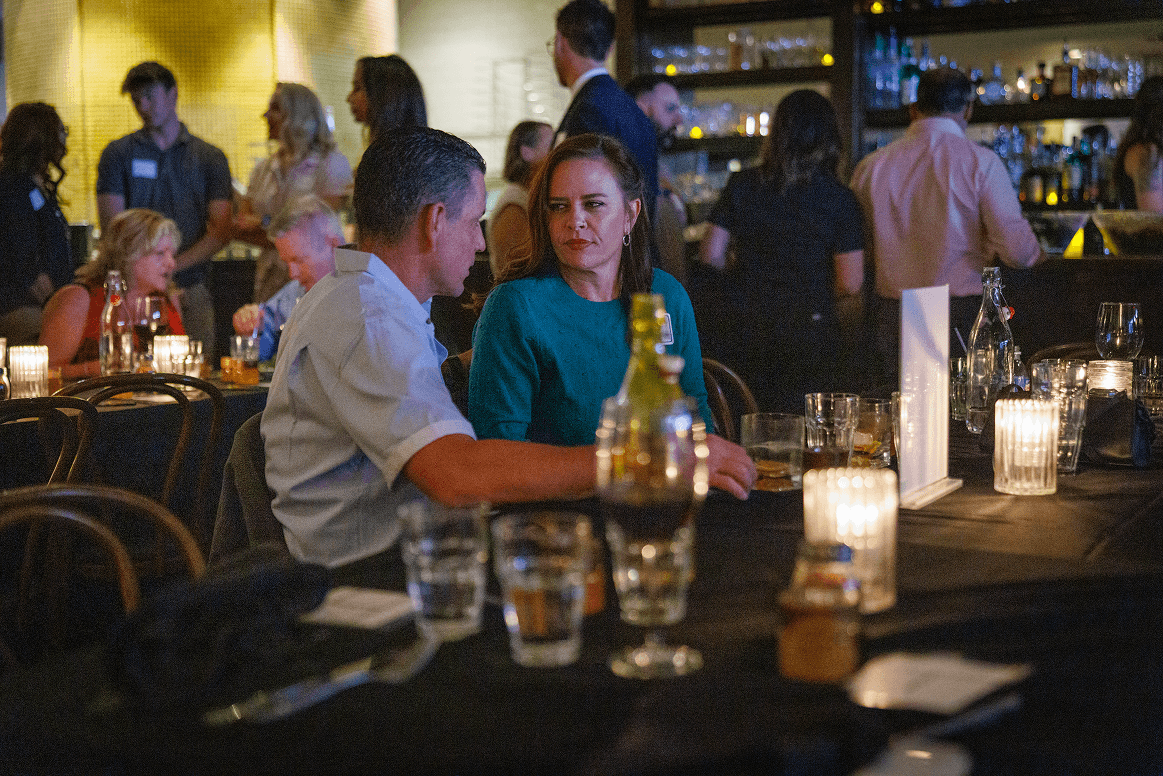 Two people, a man and a woman, converse at a candlelit table during a social event in a dimly lit restaurant with other attendees and a bar in the background.