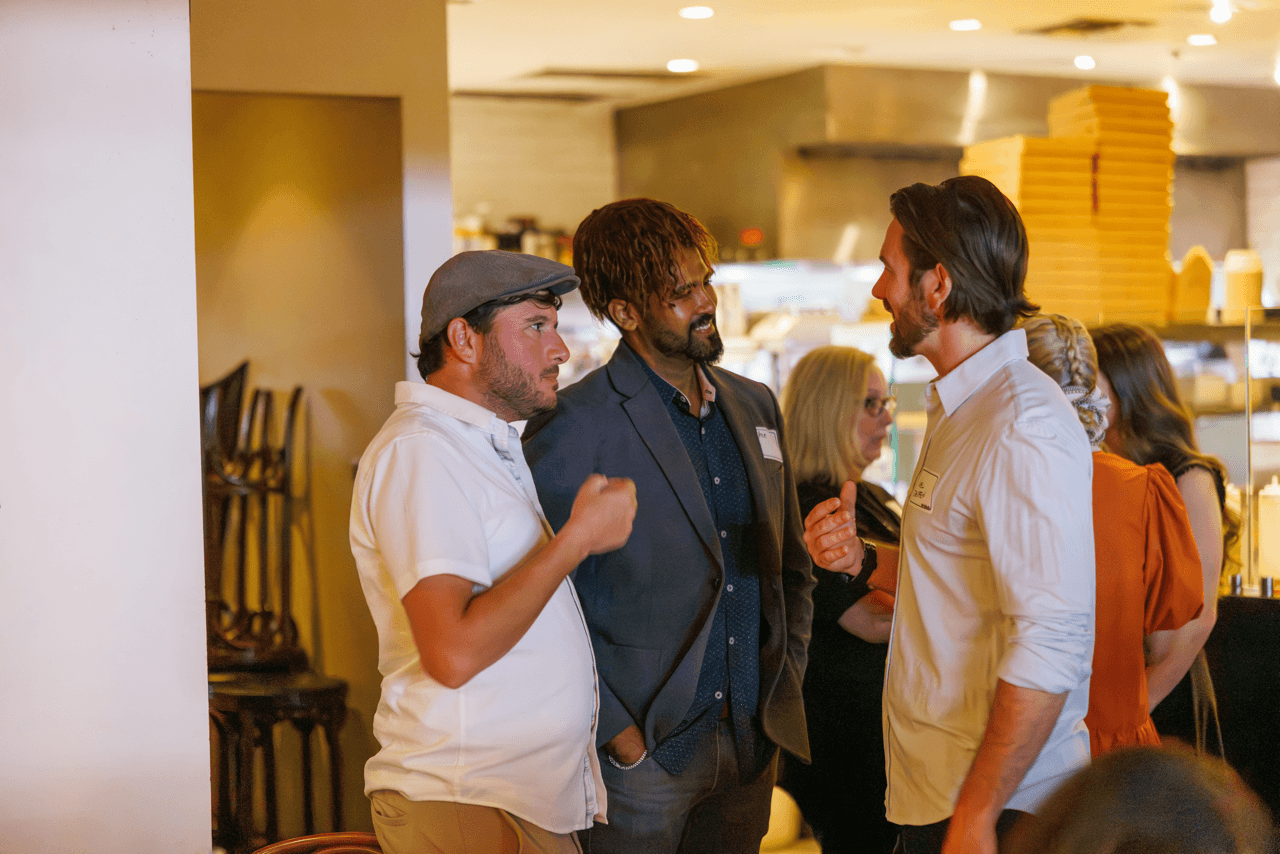 Three men engage in conversation at an indoor social event, with other attendees and a kitchen setting visible in the background.