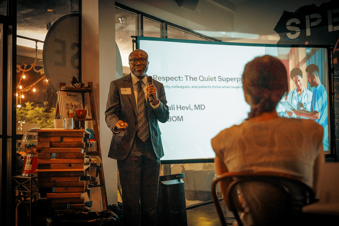 Kuali Hevi, MD, in a suit, speaks into a microphone while presenting "Respect: The Quiet Superpower" on a large screen to an audience.