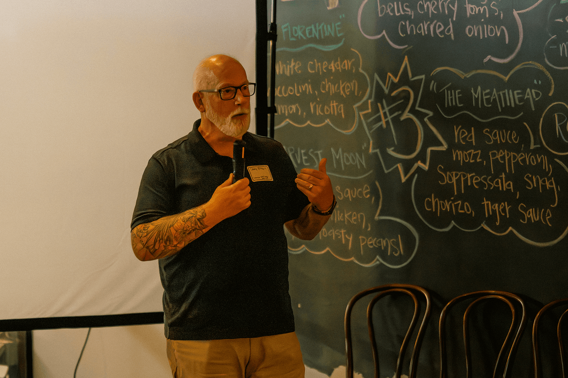 A bearded man in glasses with tattooed arms speaks into a microphone and gestures. A blank projection screen is on his left, and a dark chalkboard with menu-like writing is on his right.