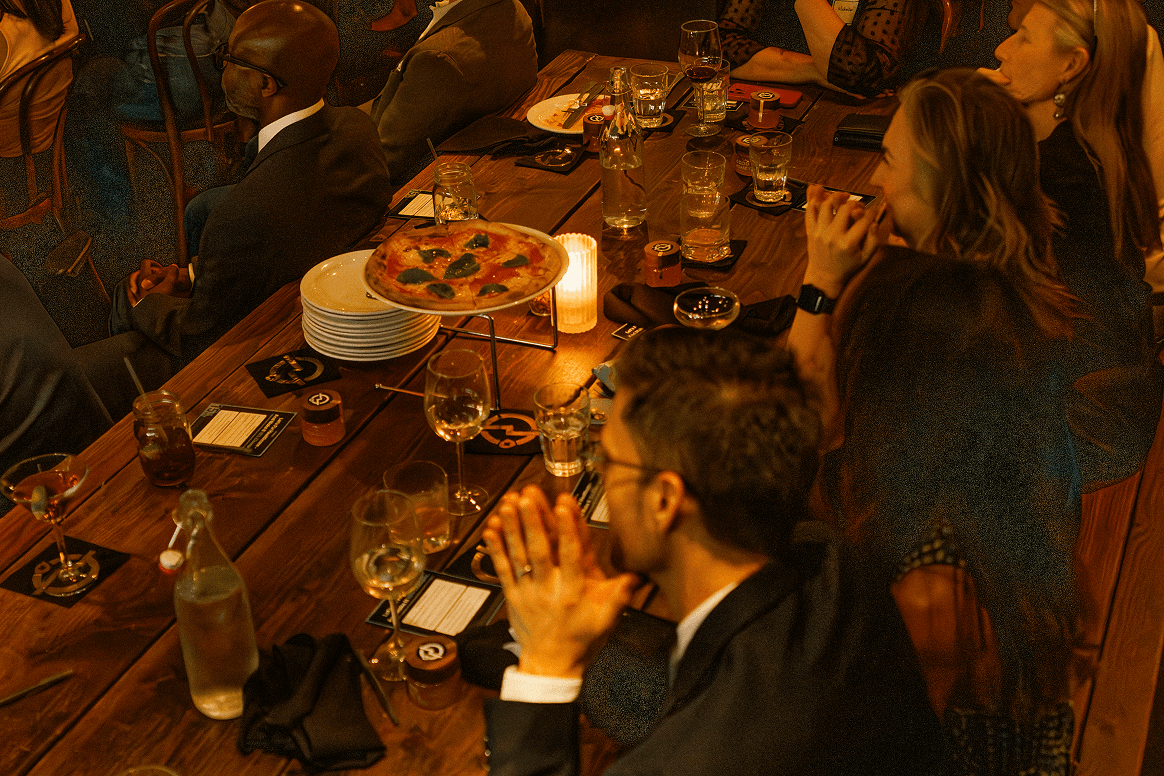 People gathered around a long wooden table with pizza, drinks, and game cards, enjoying a social event.