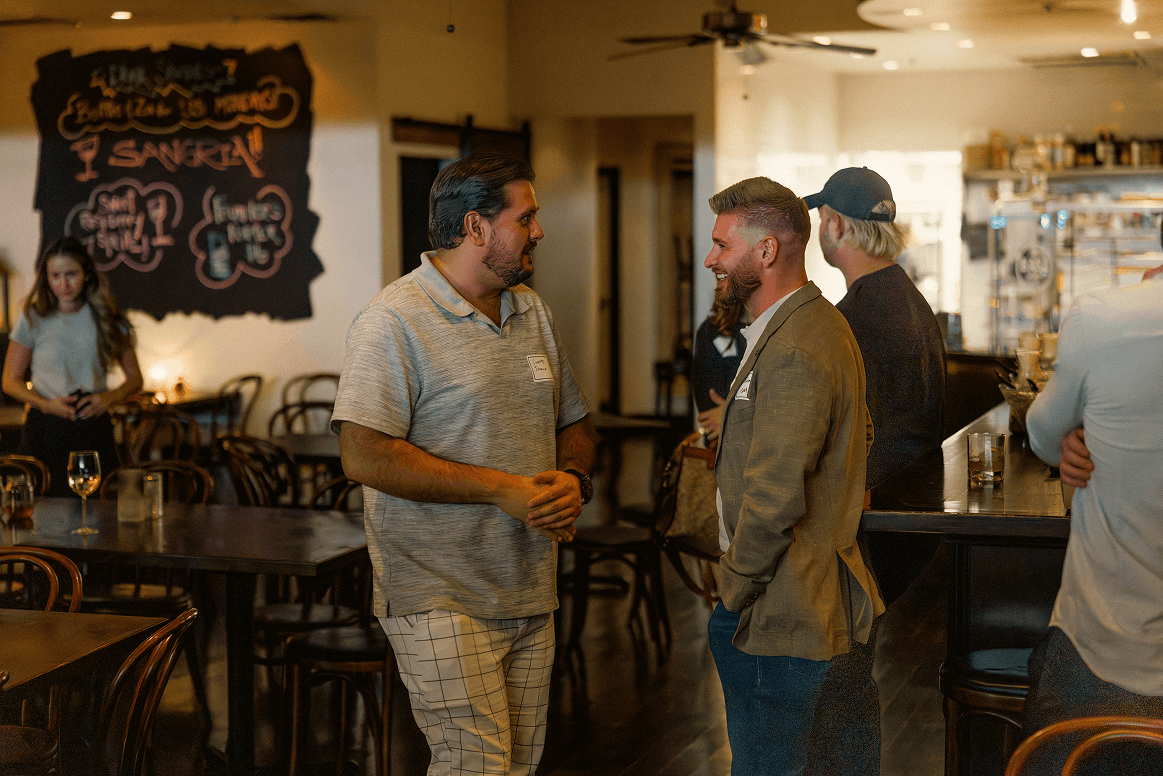 Two men with name tags are smiling and talking at a professional or social event in a dimly lit restaurant, with a blackboard menu and bar in the background.