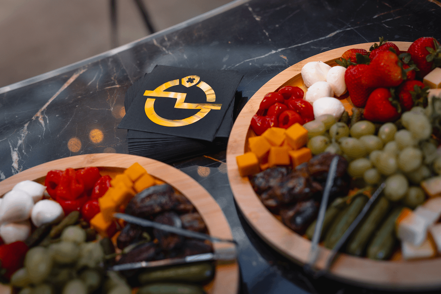 Two wooden platters laden with snacks including cheese, grapes, strawberries, and dates, next to a stack of black napkins featuring a gold logo with a stylized lightning bolt and medical cross.