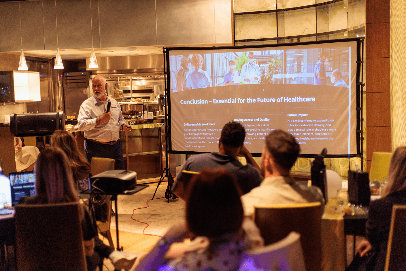 A male speaker presents "Conclusion - Essential for the Future of Healthcare" on a large screen to an attentive audience.