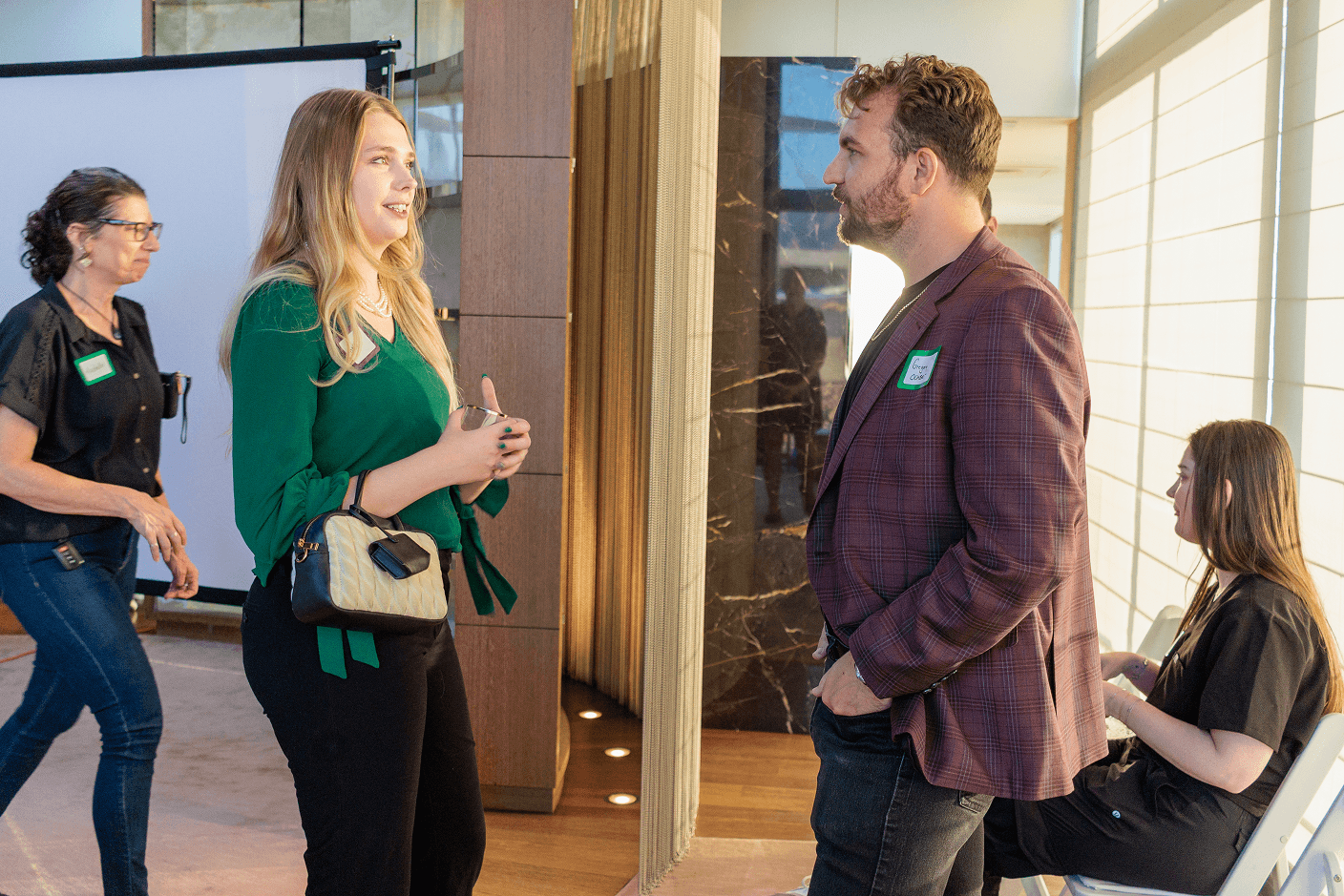 A blonde woman in a green shirt animatedly talks to a bearded man in a plaid jacket at a professional event, with other attendees nearby.