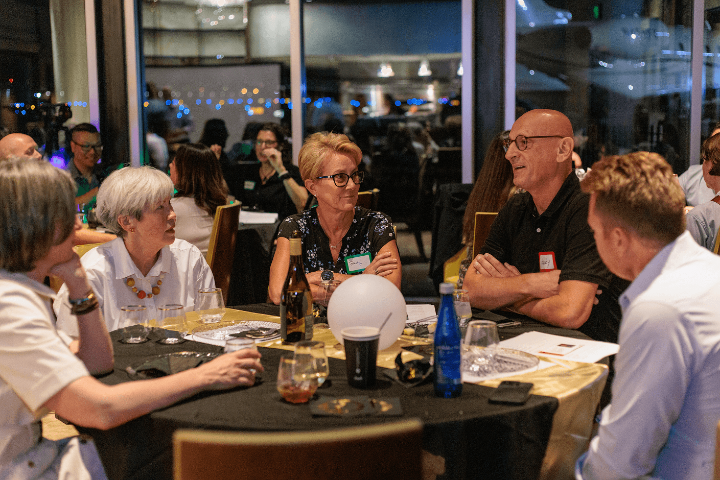 A group of professionals, some with name tags, engaged in conversation around a table at a nighttime event, with city lights visible in the background.