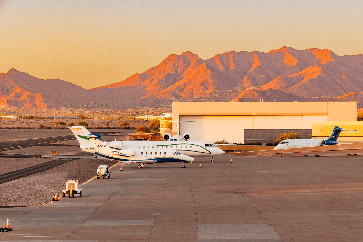 Three private jets parked on an airport tarmac, bathed in the warm light of a sunset, with sun-drenched mountains in the background.