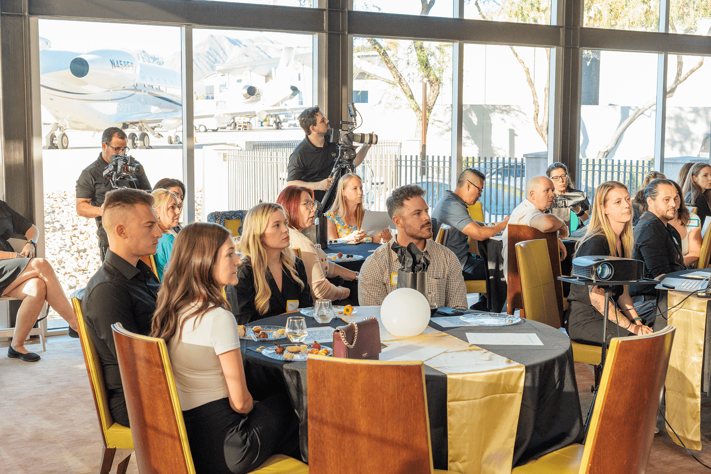 A group of professionals seated at tables during an event, with videographers filming in the background and private jets visible outside large windows.