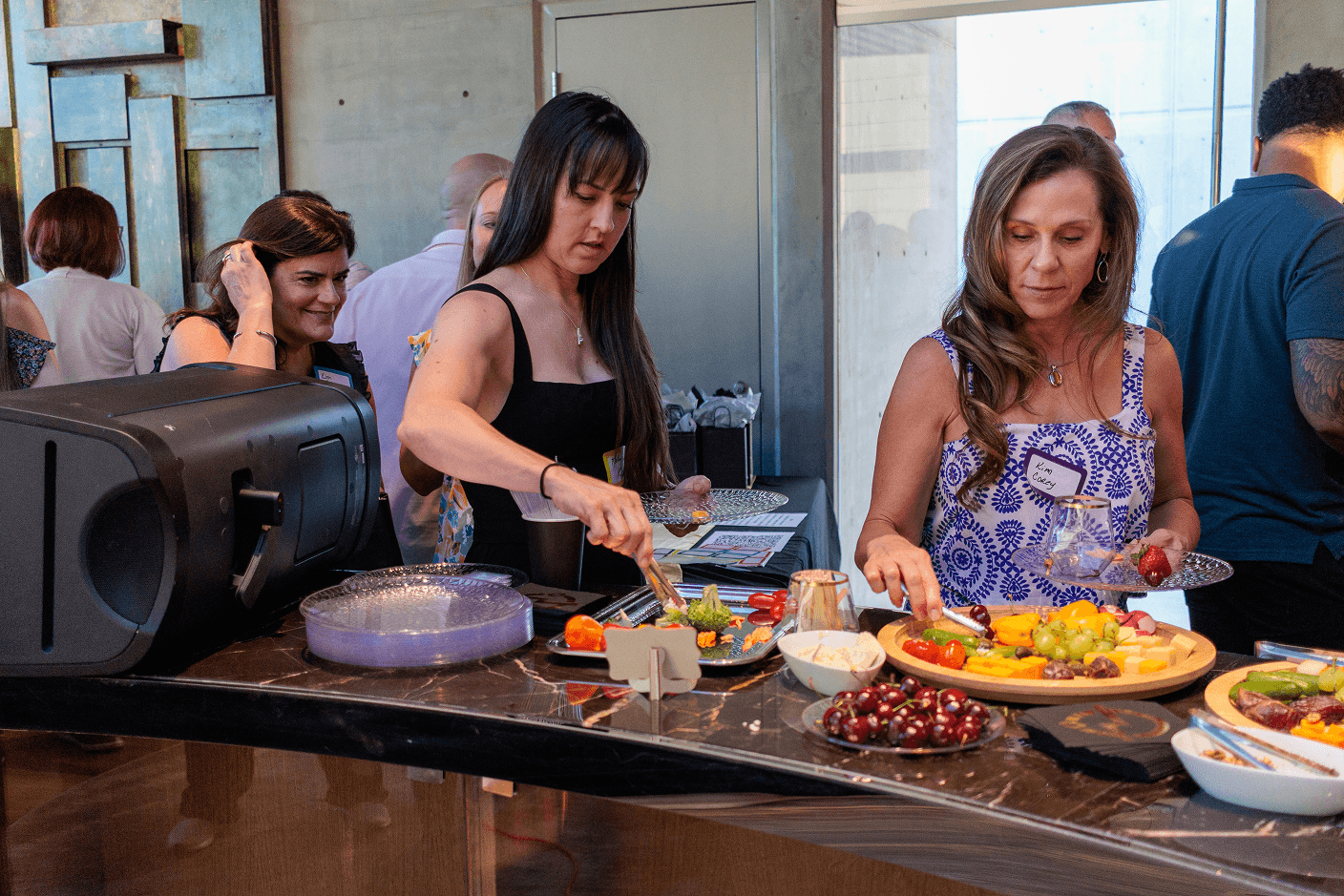 Two women serve themselves appetizers and fruit from a buffet at an indoor event, with other guests visible in the background.