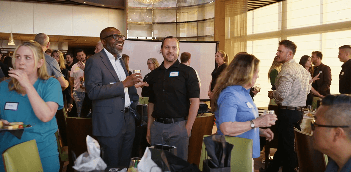 A diverse group of people networking at an indoor event. A man in a suit laughs, while a man in a black shirt smiles towards the camera. Others mingle in the well-lit room, some holding drinks or food.
