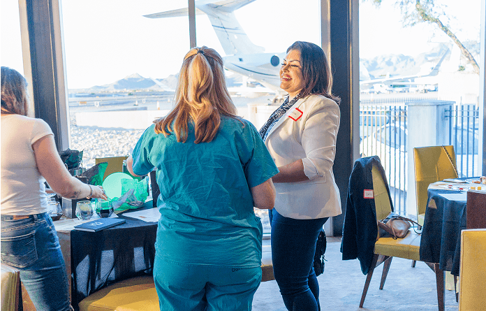 Two women, one in a white blazer smiling at another in teal scrubs, interact at a table with an airport runway and mountains visible through the large window.